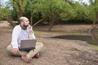 Adam dışarıda oturuyor, doğada, dizüstü bilgisayarıyla çalışıyor. O, manzarayı düşünürken düşünüyor. Konsept: dijital göçebe seyahat ederken uzaktan çalışıyor, özgürlüğün tadını çıkarıyor.