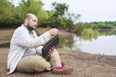 Adam bir süre akarsu kenarında çalışmak için bilgisayarını çıkarıyor. Konsept: dijital göçebe seyahat ederken uzaktan çalışıyor, özgürlüğün tadını çıkarıyor.