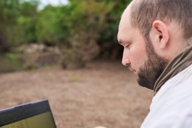 Hispanik bir adam dışarıda oturuyor, doğada, dizüstü bilgisayarıyla çalışıyor. Konsept: dijital göçebe seyahat ederken uzaktan çalışıyor, özgürlüğün tadını çıkarıyor.