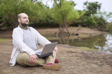 İspanyol adam dışarıda oturuyor, dizüstü bilgisayarı üzerinde çalışırken gülümsüyor. Konsept: dijital göçebe seyahat ederken uzaktan çalışıyor, özgürlüğün ve doğayla temasın tadını çıkarıyor.