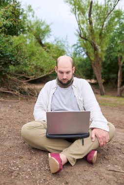 Hispanik bir adam dışarıda oturuyor, doğada, dizüstü bilgisayarıyla çalışıyor. Konsept: dijital göçebe seyahat ederken uzaktan çalışıyor, özgürlüğün tadını çıkarıyor.