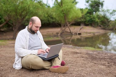 Hispanik bir adam dışarıda oturuyor, doğada, dizüstü bilgisayarıyla çalışıyor. Konsept: dijital göçebe seyahat ederken uzaktan çalışıyor, özgürlüğün tadını çıkarıyor.