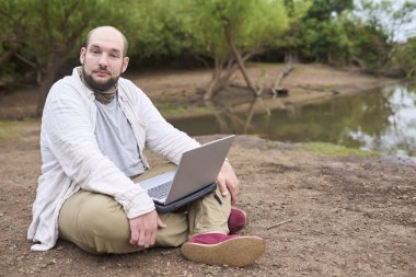 Hispanik bir adam dışarıda oturuyor, doğada, dizüstü bilgisayarıyla çalışıyor. Konsept: dijital göçebe seyahat ederken uzaktan çalışıyor, özgürlüğün tadını çıkarıyor.