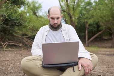 Hispanik bir adam dışarıda oturuyor, doğada, dizüstü bilgisayarıyla çalışıyor. Konsept: dijital göçebe seyahat ederken uzaktan çalışıyor, özgürlüğün tadını çıkarıyor.