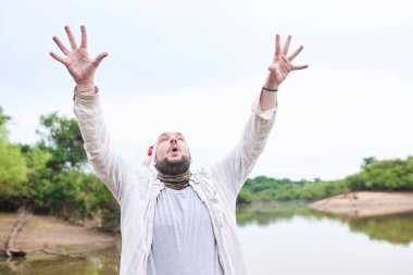 Doğal bir manzarada Noel şapkası takan adam, iki kolu havada ve yukarı bakıyor..