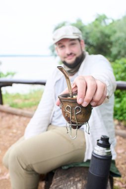 Gülümseyen adam kameraya eş teklif ediyor. Bu Arjantin içkisi paylaşımın ve birlikteliğin sembolüdür. Seçici bir şekilde ele odaklı kompozisyon.