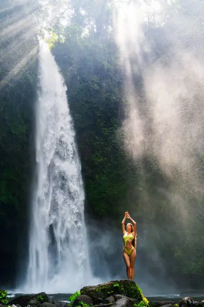 stock image Woman in yellow swimsuit poses on a rock by a sunlit waterfall.