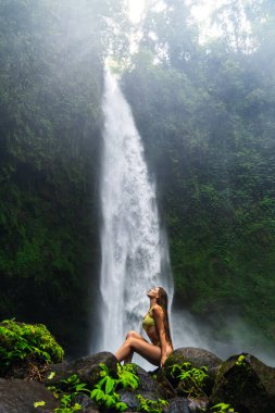 Açık yeşil mayo giymiş bir kadın güçlü bir şelalenin önünde rahat ve neşeli bir şekilde kayanın üzerinde oturuyor..