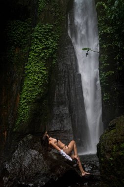 Siyah bikinili ve beyaz gömlekli bir kadın, arka planında uzun bir şelale bulunan yemyeşil kayaların üzerinde dramatik bir şekilde poz veriyor..