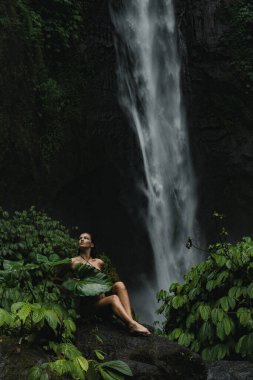 Şelalenin önünde büyük bir yaprak ile poz veren kadın, orman yapraklarıyla çevrili..