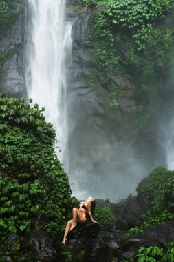 Şelalenin yakınındaki kayalarda oturan bikinili bir kadın, yemyeşil ormanlarla çevrili.