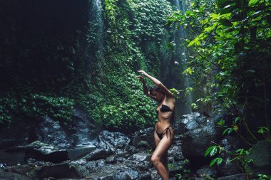 Siyah bikinili bir kadın, küçük bir şelalenin yanındaki yemyeşil tropikal bir yerde kollarını kaldırır..
