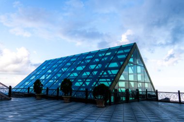 Parlak gökyüzünün altında, saksı bitkileri ve süslü çitlerle çevrili, geometrik dizaynlı modern mavi cam bina..