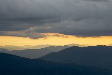 Gün batımında dramatik bir gökyüzünün altındaki katmanlı dağların nefes kesici manzarası. Işığın ve gölgenin etkileşimi çarpıcı, sakin bir manzara yaratır..