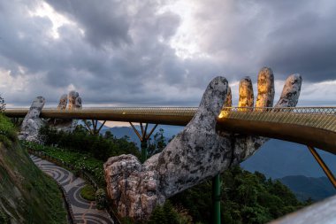 Vietnam 'daki Altın Köprü, gece aydınlanır ve dev taş ellerle desteklenir bulutlu gökyüzüne karşı çarpıcı ve dramatik bir sahne yaratır. Çevreleyen yeşillik büyülü atmosferi güçlendirir..