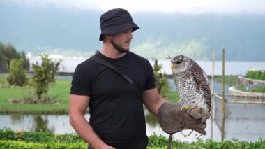 Siyah kıyafetli ve kova şapkalı bir adam eldivenli elinde baykuş tutuyor. Sahne yeşillik ve dağlarla dolu güzel bir arkaplan üzerine kurulmuş..