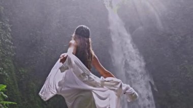 Siyah giysili ve şapkalı bir kadın, elinde beyaz bir bezle görkemli bir şelalenin yanındaki yağmuru neşeyle kucaklıyor. Yeşillik ve güçlü Nungnung şelalesi çarpıcı bir doğal manzara yaratır..