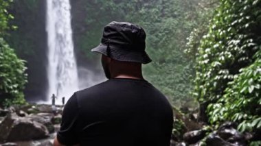 Üzerinde kelebek olan siyah bir kostüm ve kova şapka giyen bir adam, yemyeşil yapraklarla çevrili Nungnung Şelalesi 'nin tadını çıkarıyor. Huzurlu doğal ortam güzelliği vurgular.