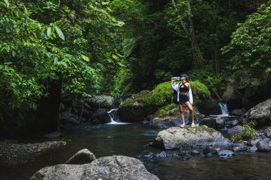 Yoğun bir ormanın arka planı ve akan bir akarsu. Bir kadın, doğanın huzurunu kucaklayarak ferahlatıcı bir içecek alır.