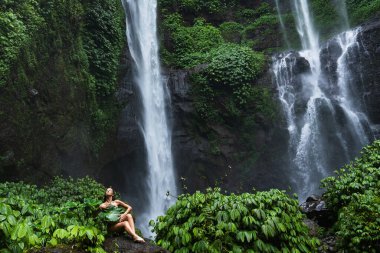 Çağlayarak çağlayan bir şelalenin önündeki kayanın üzerinde oturan ve yemyeşil çimlerle çevrili büyük bir yaprak tutan kadın..