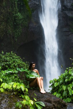 Bikinili bir kadın şelalenin yakınındaki bir kayanın üzerinde dinleniyor ve arka planda sık orman yaprakları var..