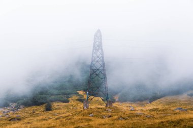 Metal bir elektrik kulesi sisli bir dağ manzarasında duruyor. Yamaçlardaki sarı çimenler ve yeşil çalılar, gökyüzünü kaplayan yoğun sis, sakin ve dengesiz bir atmosfer yaratıyor..