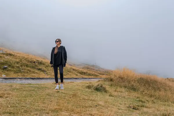 stock image A young, beautiful woman in black stands by the roadside in the mountains, with dense fog behind her.