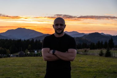 Kırsal bir alanda kollarını kavuşturmuş duran bir adam, canlı bir gün batımı ve arka planda dağlarla.