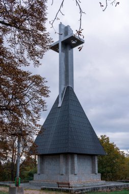 Büyük bir metal haç heykeli, bulutlu bir gökyüzünün altında, sonbahar ağaçlarıyla çevrili taştan bir üste duruyor..