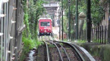 Kırmızı füniküler bir tren, bereketli yeşilliklerle çevrili, dik bir yolda yol alır ve teknoloji ile doğa arasında bir zıtlık yaratır. Eşsiz demiryolu taşımacılığının cazibesini yakalar.