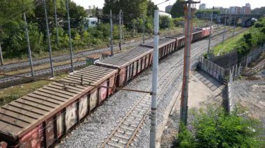 Kırmızı bir yük treni, yeşil ve endüstriyel unsurlarla çevrili işlek bir kentsel çevrede, bir şehir manzarasında ulaşım ve hareketi tasvir eden birden fazla demiryolu hattı boyunca seyahat eder..