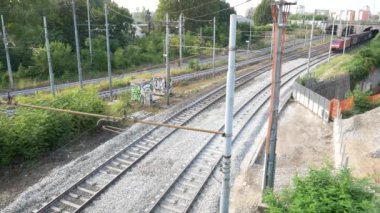Bir kırmızı yük treni, yemyeşil ve şehir manzarası ile çevrili şehir demiryolu rayları boyunca hızla seyahat eder. Enerji ve hareket modern ulaşımın özünü yakalıyor..