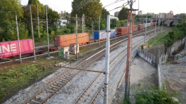 Bir kargo treni yeşil ve kentsel unsurlarla çevrili demiryolu rayları boyunca hızla hareket eder. Sahne ulaşım ve endüstriyel hareketin özünü yakalıyor..