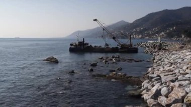 Camogli 'nin kayalık kıyı şeridini, kenetlenmiş bir gemiyi ve uzak dağları gösteren manzaralı bir kıyı manzarası. Huzurlu deniz manzarası bir huzur ve macera hissi yakalar..