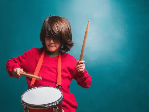 Okul tahtasının önünde davul çalan kırmızı kazaklı bir çocuk.