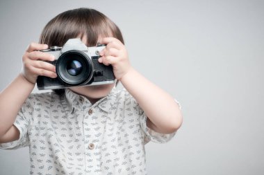 Beyaz gömlekli ve mavi elbiseli bir çocuk antika fotoğraf makinesiyle fotoğraf çekiyor..  