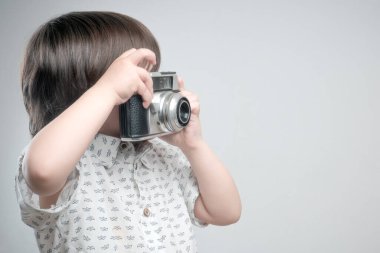 Beyaz gömlekli ve mavi elbiseli bir çocuk antika fotoğraf makinesiyle fotoğraf çekiyor..  