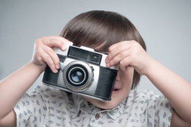 Beyaz gömlekli ve mavi elbiseli bir çocuk antika fotoğraf makinesiyle fotoğraf çekiyor..  