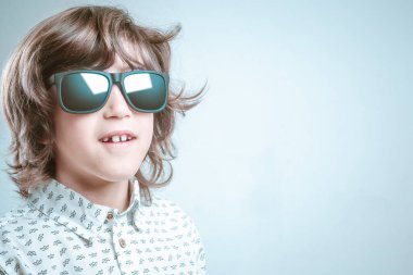 Beyaz gömlekli ve mavi elbiseli bir çocuk fotoğraf çekiminde güneş gözlüklü poz veriyor..