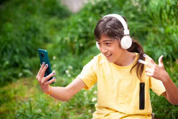 Çocuk cep telefonundan beyaz kulaklıklarla müzik dinliyor ve baharda yeşil bir çayırın yanında selfie çekiyor.. 