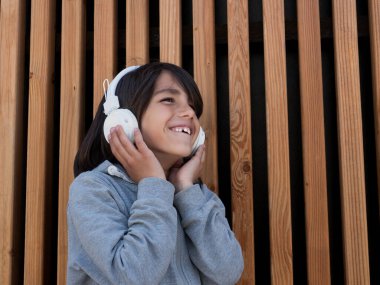 Çocuk ahşap bir duvarın önünde beyaz kulaklıkla müzik dinliyor.. 