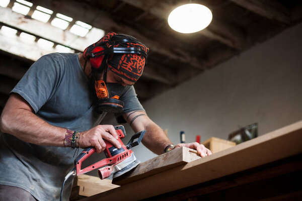 In the bustling workshop, a skilled woodworker is captured polishing a board with a random orbital sander. The image showcases the artisan's craftsmanship, while offering abundant copy space for personalized messages or branding.