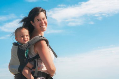 Anne, çocuğunu sırtında bir sırt çantasıyla ve yüzünde kocaman bir gülümsemeyle taşıyor..