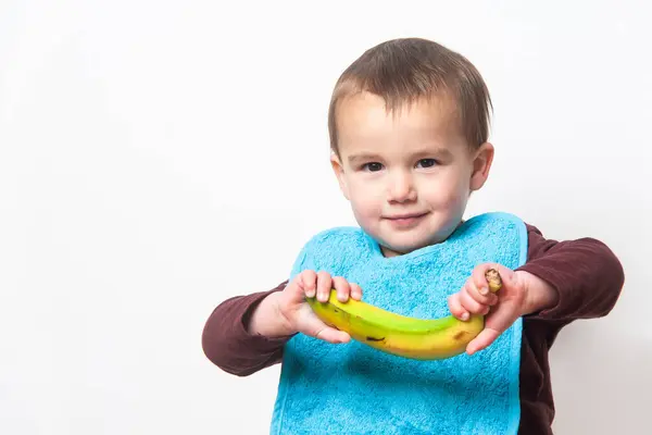 Çocuk elinde muz tutarken kameraya bakıyor. Kirlenmemek için mavi önlük giyiyor. Uzayı kopyala Beyaz arka plan. İzole edilmiş.