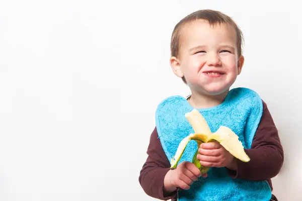 Mavi önlüklü çocuk yüzünde kocaman bir gülümsemeyle muz yiyor ve lezzetli tropikal meyvelerin tadını çıkarıyor. Uzayı kopyala Beyaz arka plan. İzole edilmiş.