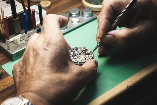 The hands of a watchmaker repairing a watch. They are manipulating a watchmaker\'s tweezers. . Other tools can be seen in the background.