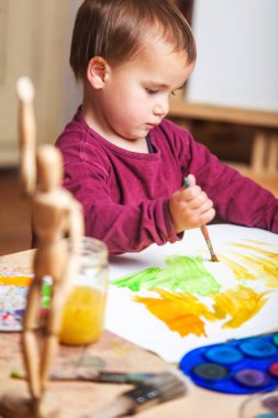 Çalışma masasındaki beyaz tuvalinde renkli akrilik boyalarla boyalı, farklı fırça, boya ve çömlek resimleri olan küçük bir çocuğun dikey görüntüsü.. 
