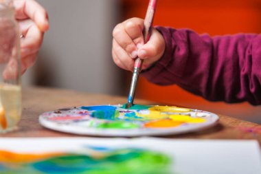 Yaratıcı çalışmalarına devam etmek için bir renk paletinden akrilik boya topladığı bir fırçayla küçük bir çocuğun elinin detaylı görüntüsü.. 