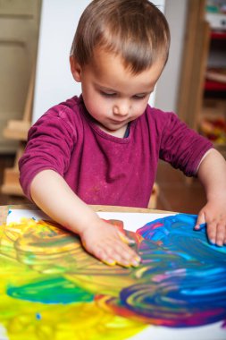 Yeni yürümeye başlayan çocuk parlak renkli akrilik boyalarla bir tuval boyamaya ve renk karışımı yaratmaya odaklanıyor.. 