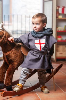 Küçük çocuk bir eliyle sallanan atını, diğer eliyle tahta kılıcını evdeki oyun odasında tutuyor..